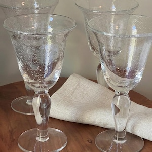 May include: Four clear, textured wine glasses with a bubble-like design. The glasses have a flared bowl, a clear stem, and a round base. A beige linen napkin rests beneath the glasses on a wooden surface.