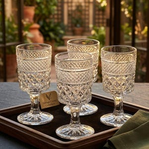 May include: Four clear glass goblets featuring a textured diamond pattern on the bowl and a short stem. A small rectangular tag attached to one glass reads Vintage Crystal Collection. The glasses are arranged on a dark wooden tray.