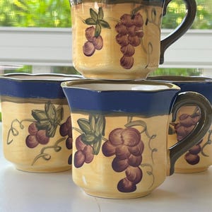 May include: Set of four ceramic mugs. Each mug features a yellow body with a painted grape design in shades of purple and green. The rim and handle are dark blue and the handle is dark green.