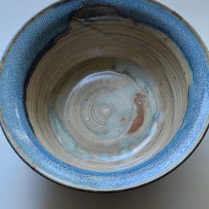 May include: A handmade ceramic bowl with a light blue rim and a beige interior. The bowl has a rustic, textured appearance with visible brushstrokes and a unique, organic shape. The interior features concentric circles.