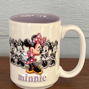 May include: Cream-colored ceramic mug featuring Minnie Mouse in a red polka-dot dress and bow. The mug has a purple interior and the word "minnie" in purple script. Several black and white Minnie Mouse figures are also on the mug.