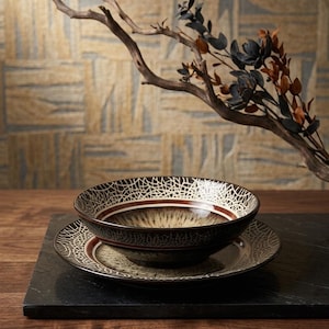 May include: A ceramic bowl and plate set with a brown and cream color scheme. The bowl has a unique white web-like pattern around the rim and a brown stripe. The plate also features the same web-like pattern. A decorative branch with leaves is in the background.