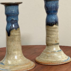 May include: Two ceramic candle holders. The top portion is glazed in a deep blue, transitioning to a tan base. The candle holders have a flared base and a cylindrical body. The glaze has a glossy finish.