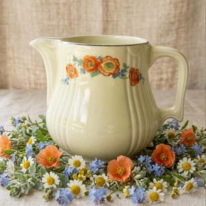 May include: A cream-colored ceramic pitcher with a vertical ribbed texture and a silver rim. The body features a painted floral pattern of orange poppies and small blue flowers. The pitcher has a curved handle and a wide pouring spout.