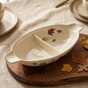 May include: Cream-coloured, divided serving dish with leaf designs. The oval dish has two compartments and handles. It sits on a dark wooden serving board, with plates and utensils in the background. Measurements in cm.