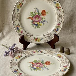 May include: Two vintage porcelain plates with floral designs. The plates have a cream-colored background with a gold rim and intricate gold filigree. Each plate features a central bouquet of pink, red, blue, and yellow flowers. Two thimbles and a small bouquet are also visible.