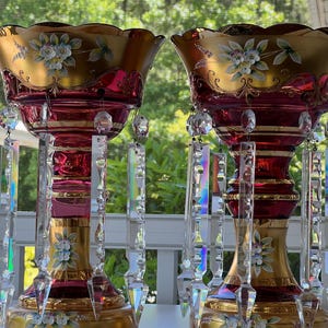 May include: Two ornate, ruby red glass vases with gold rims and bases. Each vase features hand-painted floral designs in white and blue, with dangling crystal accents. The vases are likely decorative items.