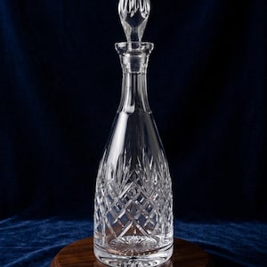 May include: A clear crystal decanter with a diamond-cut pattern and a teardrop-shaped stopper. The decanter sits on a round, dark wooden base against a dark blue background. The stopper is ribbed and tapers to a point.