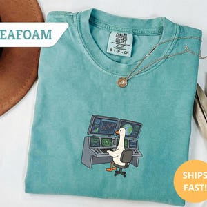 May include: Seafoam green Comfort Colors t-shirt with a graphic of a goose sitting at a computer desk with two monitors. The monitors display charts and graphs. The shirt has a small tag and a gold necklace. The words "SHIPS FAST!" are in a circle.