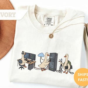 May include: Ivory-colored t-shirt with a graphic of cartoon geese in a tech-related theme. One goose is at a computer, another with a cloud, and the third with cables and a keyboard. The shirt has a Comfort Colors tag.