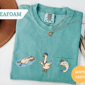 May include: Seafoam green t-shirt with three cartoon geese in swimming poses. One goose floats on a float, another gives a thumbs up with a medal, and the third dives into the water. A gold necklace is draped on the shirt.
