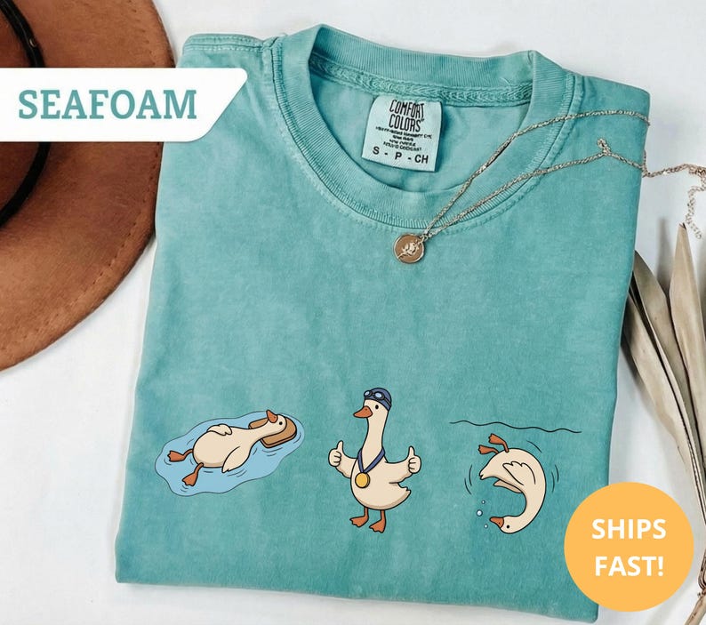May include: Seafoam green t-shirt with three cartoon geese in swimming poses. One goose floats on a float, another gives a thumbs up with a medal, and the third dives into the water. A gold necklace is draped on the shirt.