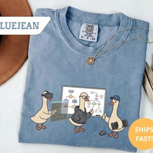 May include: Blue jean colored t-shirt with a graphic of three cartoon geese in a tech-related theme. One goose wears a VR headset, another wears glasses and a hoodie, and the third wears a cap and holds a keyboard. The shirt has the text "BLUEJEAN".