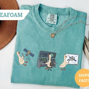 May include: Seafoam green t-shirt with a cartoon goose theme. The shirt features three illustrations of geese working on a satellite. The text "SHIPS FAST!" is on a yellow circle.