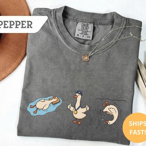 May include: A gray Comfort Colors t-shirt featuring three cartoon geese in swimming-themed poses. One goose floats on a pool float, another gives a thumbs-up with a medal, and the third dives into water. The shirt has the word "PEPPER" and "SHIPS FAST!" on it.