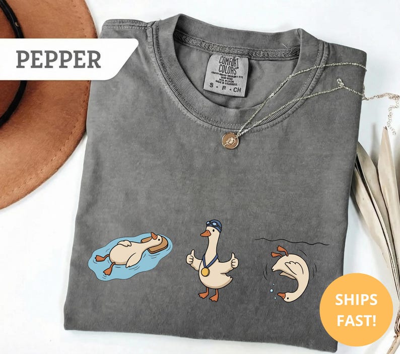 May include: A gray Comfort Colors t-shirt featuring three cartoon geese in swimming-themed poses. One goose floats on a pool float, another gives a thumbs-up with a medal, and the third dives into water. The shirt has the word "PEPPER" and "SHIPS FAST!" on it.
