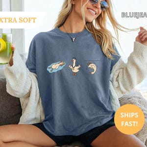 May include: Blue jean colored t-shirt with a cartoon goose design. The goose is depicted in three different poses: floating, standing, and diving. The shirt has the text "EXTRA SOFT" and "BLUEJEAN".