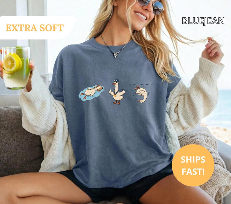 May include: Blue jean colored t-shirt with a cartoon goose design. The goose is depicted in three different poses: floating, standing, and diving. The shirt has the text "EXTRA SOFT" and "BLUEJEAN".