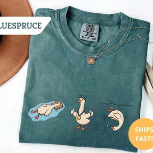 May include: Teal Comfort Colors t-shirt featuring three cartoon geese in swimming poses. One goose floats on a raft, another gives a thumbs up with a medal, and the third dives into the water. The shirt has the text "BLUESPRUCE" and "SHIPS FAST!"
