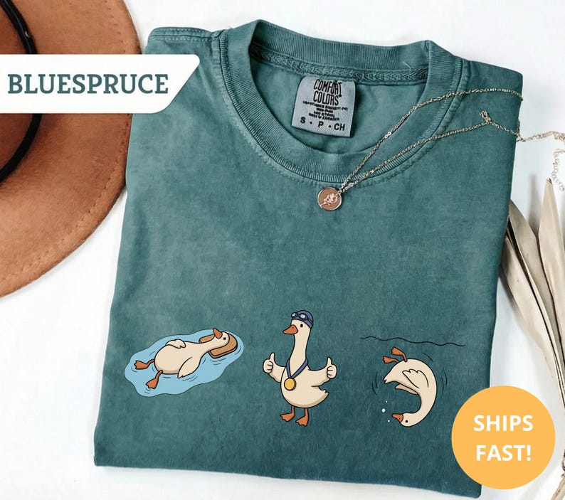 May include: Teal Comfort Colors t-shirt featuring three cartoon geese in swimming poses. One goose floats on a raft, another gives a thumbs up with a medal, and the third dives into the water. The shirt has the text "BLUESPRUCE" and "SHIPS FAST!"