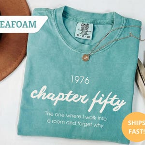 May include: Seafoam green t-shirt with the text "1976 chapter fifty" in white script. The shirt also has the phrase "The one where I walk into a room and forget why." A gold necklace with a pendant rests on the shirt.