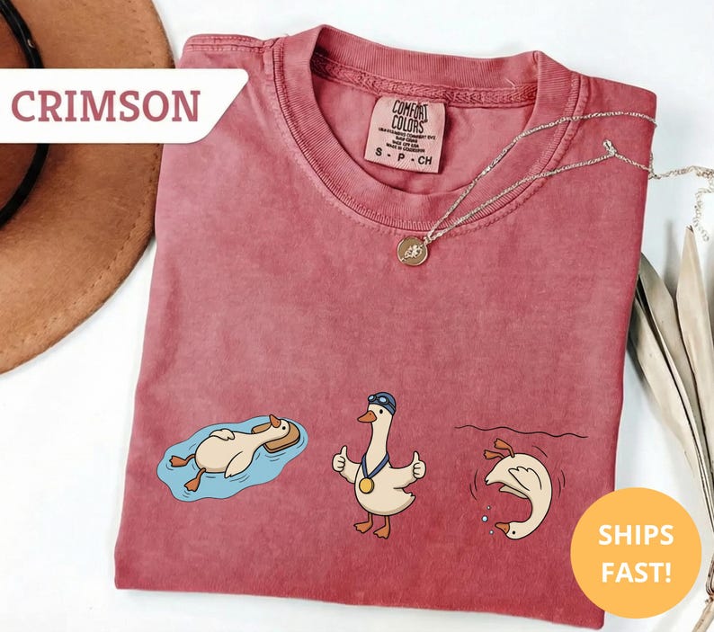 May include: Crimson-colored t-shirt with three cartoon geese in different swimming poses. One goose floats on a tube, another gives a thumbs-up with a medal, and the third dives into the water. The shirt has the text "Comfort Colors" on the tag.