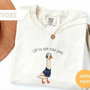 May include: Ivory-colored t-shirt with the text "Off to calm total panic" and a cartoon goose wearing a headset and a navy blue jacket. A gold necklace and a round yellow sticker are also visible.