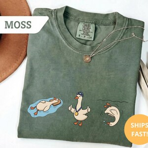 May include: Moss green t-shirt with three cartoon geese in different swimming poses. One goose is floating on a raft, another is giving a thumbs up, and the third is diving. The shirt has a Comfort Colors tag.