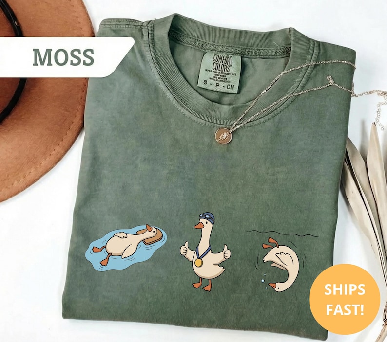 May include: Moss green t-shirt with three cartoon geese in different swimming poses. One goose is floating on a raft, another is giving a thumbs up, and the third is diving. The shirt has a Comfort Colors tag.