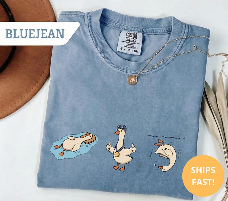 May include: A blue t-shirt with three cartoon geese in different swimming poses. One goose is floating on a float, another is giving a thumbs up, and the third is diving. The shirt has the text "BLUEJEAN" and "SHIPS FAST!"