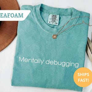 May include: Seafoam green t-shirt with the phrase "Mentally debugging" in white. A gold necklace with a pendant rests on the shirt. The shirt is a Comfort Colors brand. A brown hat is in the background.