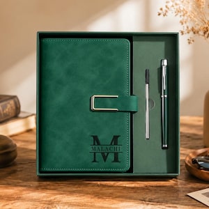 Pode incluir: Um caderno verde-escuro com um fecho retangular dourado e o nome Malachi impresso sobre uma letra M grande. O caderno está numa caixa de presente verde a condizer, juntamente com uma caneta verde-escura e uma recarga de tinta prateada.