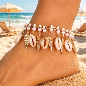 May include: Gold-toned anklet featuring a chain design with white pearls, small seashells, and gold-colored leaf charms. The anklet is worn on a foot on a sandy beach, with the ocean and beach umbrellas in the background.