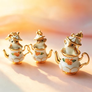Könnte beinhalten: Drei Miniatur-Teekannen-Anhänger. Jeder Anhänger zeigt eine kleine, hellbraune Maus mit weißem Hut, die in einer weißen Teekanne mit goldenen Verzierungen und orangefarbenen Blumenmotiven sitzt. Die Anhänger sind mit aufwendigen Details gestaltet.