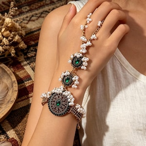 May include: A gold-toned hand chain bracelet featuring three circular filigree medallions with central emerald green stones. The piece is adorned with small white faux pearls and connects from a wrist cuff to a ring via a delicate chain.