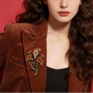 Puede incluir: Un broche floral detallado con acentos marrones, dorados y verdes, colocado sobre una chaqueta de pana marrón. El diseño floral presenta un centro de cristales brillantes y detalles de pétalos.