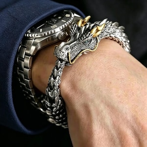 以下が含まれることがあります： 金色のアクセントが付いた銀色のドラゴンブレスレットが手首に巻かれ、銀色の腕時計の隣にあります。ドラゴンの頭には青い目と金の角があります。ブレスレットのチェーンは、連動する銀色の鱗でできています。