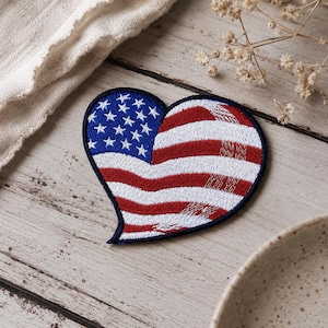 May include: A heart-shaped embroidered patch featuring the American flag design. The patch has a dark blue field with white stars on the upper left side and horizontal red and white stripes filling the remainder of the heart shape, bordered by a dark blue edge.