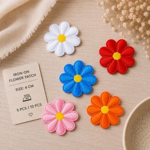Può includere: Cinque toppe ricamate a forma di fiore nei colori bianco, rosso, blu, rosa e arancione, ciascuna con un centro giallo. Una scheda rettangolare riporta il testo IRON-ON FLOWER PATCH, SIZE: 4 CM, 5 PCS / 10 PCS e un'icona di ferro da stiro.