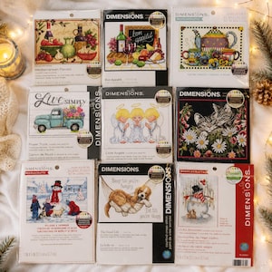 Puede incluir: Una colección de nueve kits de punto de cruz Dimensions con varios diseños, incluyendo una escena de comida mediterránea, un camión con flores, tres ángeles, un gato entre margaritas, una escena de muñeco de nieve y un perro en una hamaca.