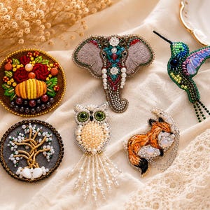 Könnte beinhalten: Eine Sammlung von sechs handgefertigten Perlenbroschen, die einen Elefanten, einen Kolibri, eine Eule, einen Fuchs, ein kreisförmiges Blumenarrangement und ein kreisförmiges Baummotiv darstellen. Jedes Stück ist mit Perlen, Pailletten und Stickereien verziert.
