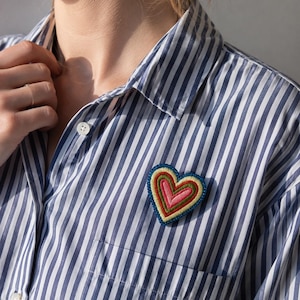 May include: A heart-shaped brooch featuring concentric layers of embroidery in shades of pink, green, yellow, and blue. The brooch is pinned to a button-down shirt with a vertical blue and white striped pattern, featuring a collar and a chest pocket.