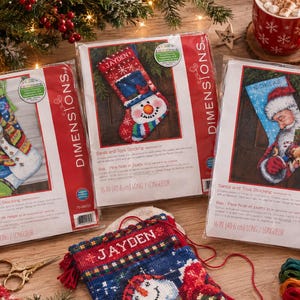 May include: Three Dimensions brand cross-stitch stocking kits in plastic packaging, featuring designs of a snowman and Santa Claus. Each kit measures 16 inches long. A partially completed stocking with the name Jayden is visible in the foreground.