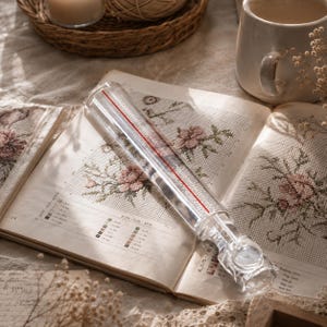 Puede incluir: Una guía de costura cilíndrica transparente con una línea roja, apoyada en un libro de patrones de punto de cruz vintage abierto. El libro presenta diseños de bordado floral en rosa y verde. Una taza y una vela están en el fondo.