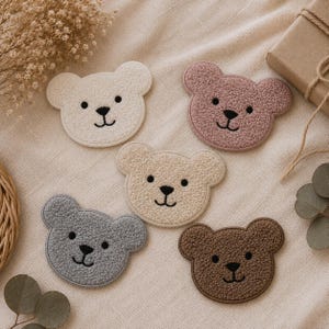 May include: Five bear-shaped patches made of textured, sherpa-like fabric in white, dusty pink, beige, gray, and brown. Each patch features embroidered black eyes, a nose, and a mouth, creating a consistent, simple facial expression across all pieces.