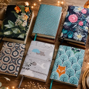 May include: Six rectangular fabric book covers featuring various patterns including dark floral prints, a teal geometric design, a grey nursery print with a zebra, and a teal forest print with a fox, each equipped with a matching ribbon bookmark.