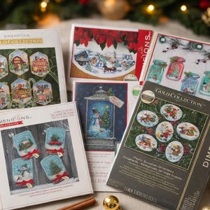Puede incluir: Una colección de kits de manualidades Dimensions Gold Collection con varios diseños de adornos navideños en punto de cruz, incluyendo muñecos de nieve, cabañas de invierno y escenas de frascos, en cajas de cartón rectangulares con imágenes.