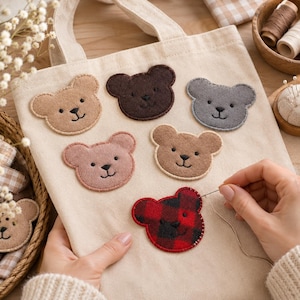 May include: A beige canvas tote bag featuring six felt bear head appliqués in various colours including tan, dark brown, grey, pink, and a red and black buffalo plaid pattern, with a needle and thread stitching the plaid bear onto the fabric.