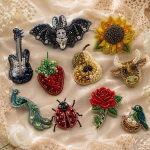 Könnte beinhalten: Eine Sammlung von Perlenbroschen in verschiedenen Formen, darunter eine Gitarre, eine Fledermaus, eine Sonnenblume, eine Erdbeere, eine Birne, eine Kuh, ein Pfau, ein Marienkäfer, eine Rose und ein Vogel im Nest. Die Broschen weisen detaillierte Perlenarbeiten in verschiedenen Farben auf.