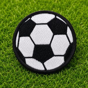May include: A circular embroidered patch featuring a classic soccer ball design with a traditional pattern of black pentagons and white hexagons. The patch has a thick black stitched border and a textured surface created by dense thread work.